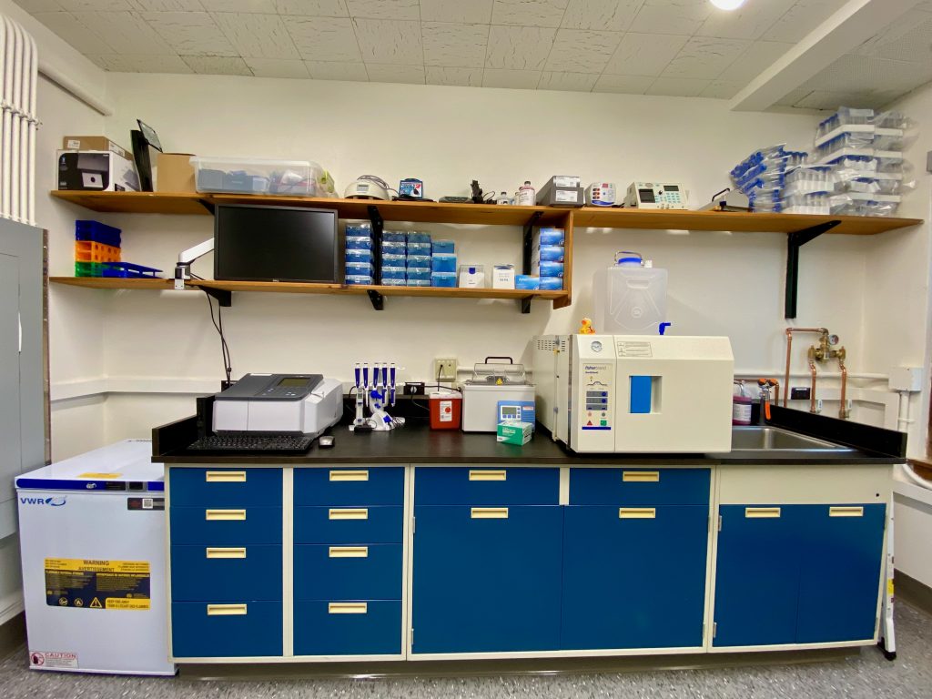 This photo depicts the workbench space in the Disinfection & Toxicology Lab, which is filled with various pieces of equipment. This includes a spectrophotometer and an autoclave. The bench top is black, and the cabinets are blue.