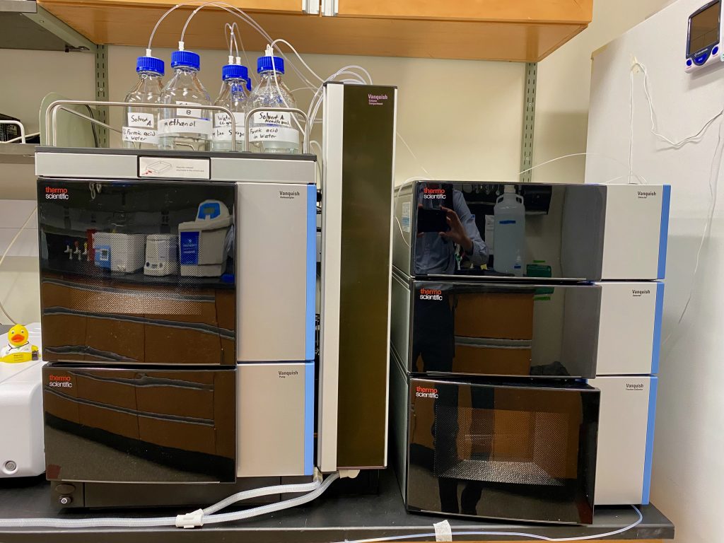The Thermo Vanquish Flex looms in this photo. It is a tall piece of scientific equipment, with black doors to the various steel-colored compartments. Bottles rest on top of the Vanquish Flex, with tubes streaming into various portions of the instrument.