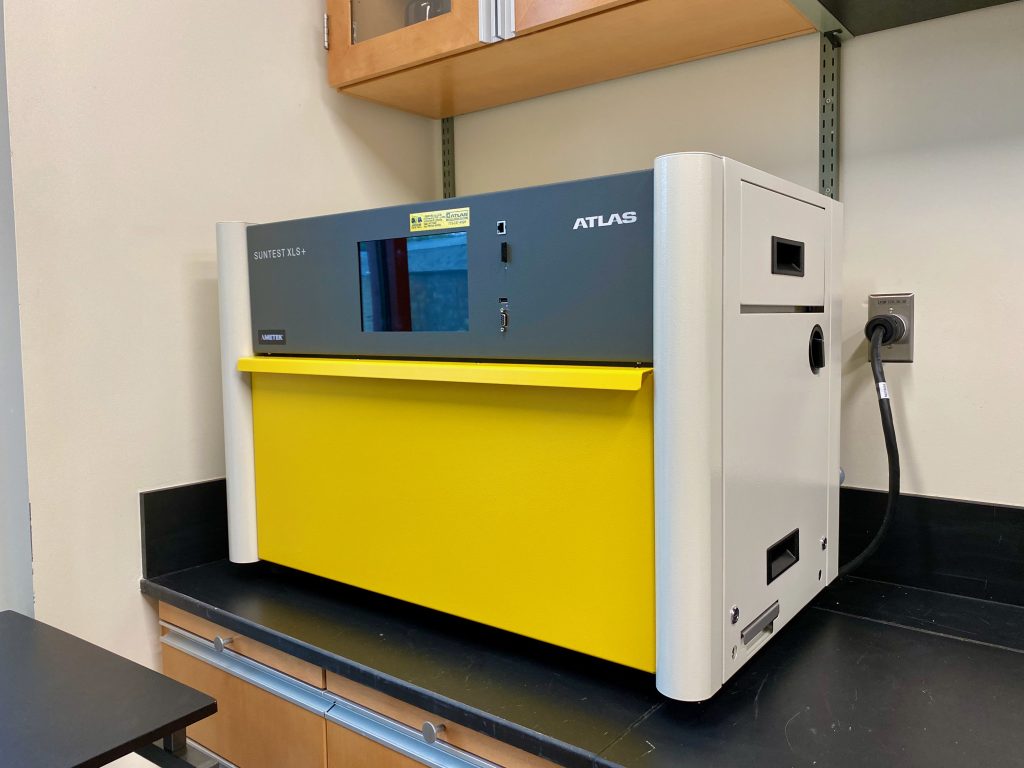 The SunTest XLS+ is a high-capacity solar simulator, allowing many samples to be irradiated simultaneously. The SunTest appears as a large aluminum box with beige sides and a large yellow compartment door that folds out toward the viewer.